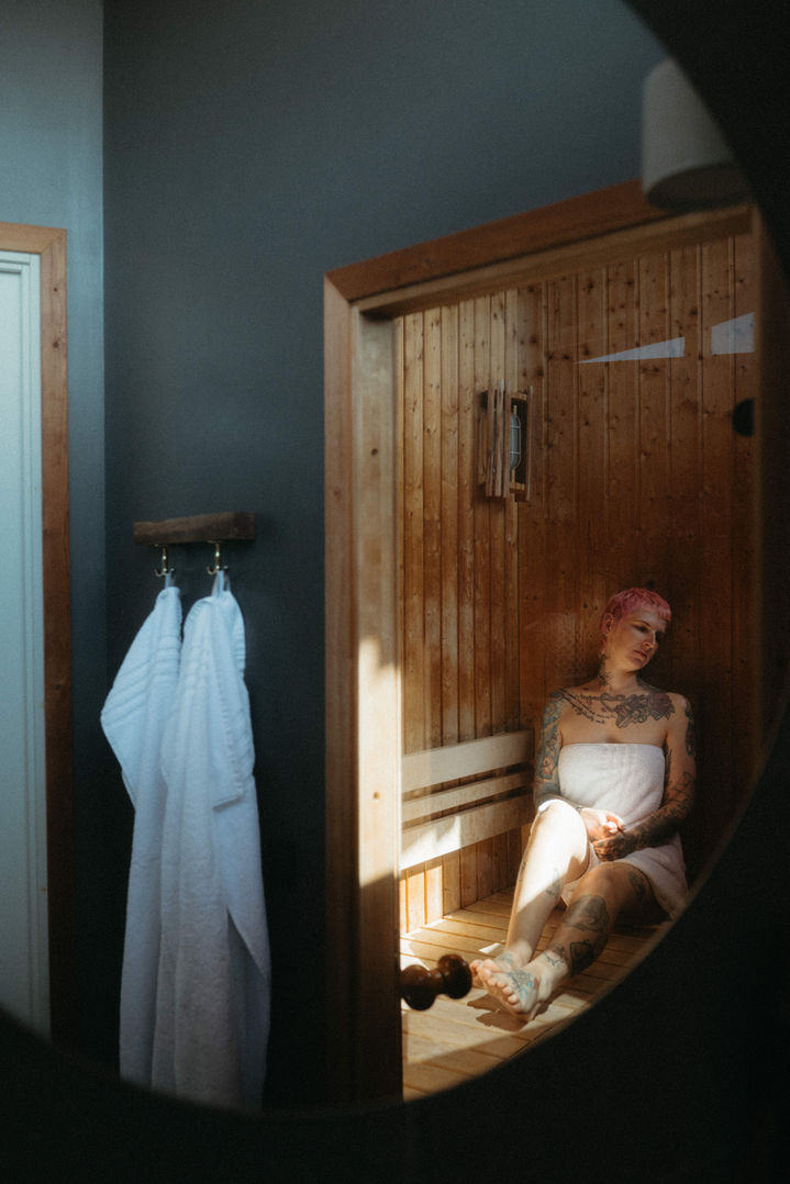 Jana sitting in a sauna at Henne Strand Resort, white robes hanging on hooks beside the wooden sauna door