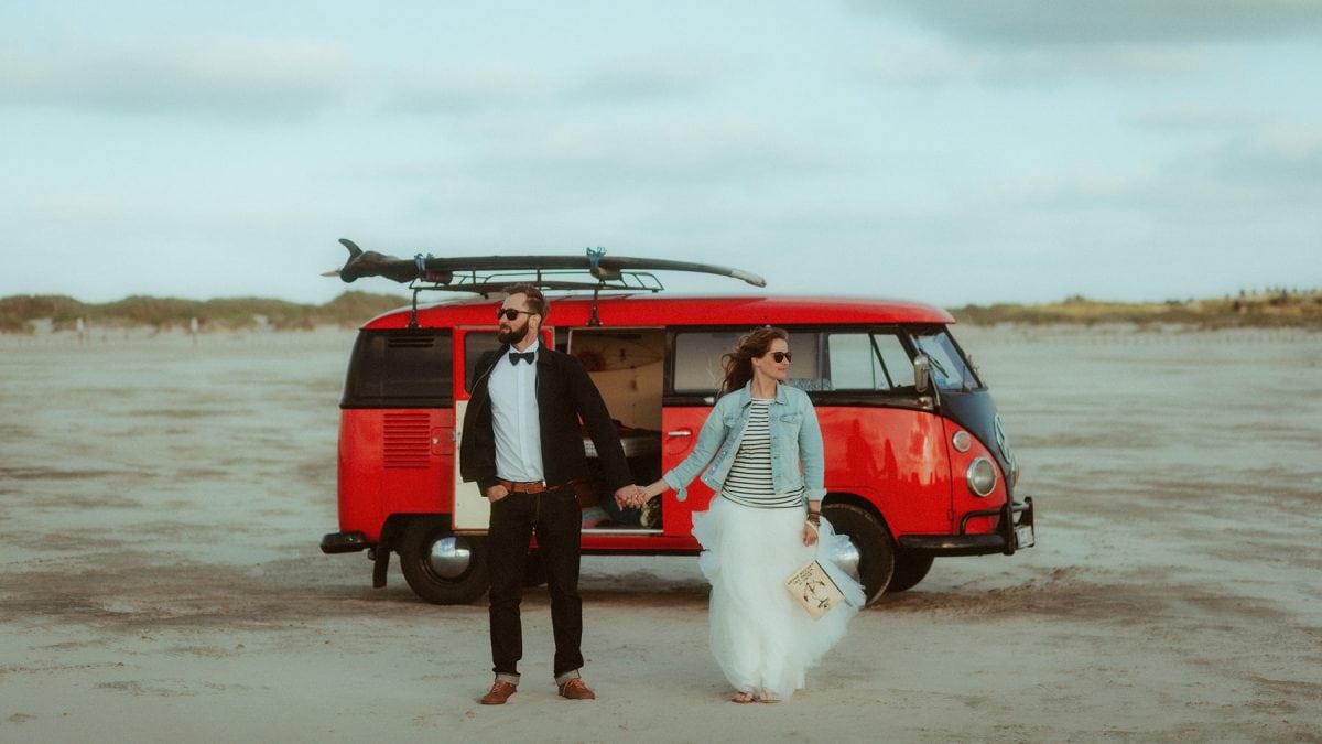 Boho-Elopement in St. Peter-Ording: Eine barfüßige Braut im Tüllrock und Jeansjacke hält die Hand ihres Partners vor einem roten Vintage-VW-Bus am weiten Nordseestrand. Lässig, windverweht und wild romantisch – nur die beiden, mit der offenen Küste als einzige Zeugin. 