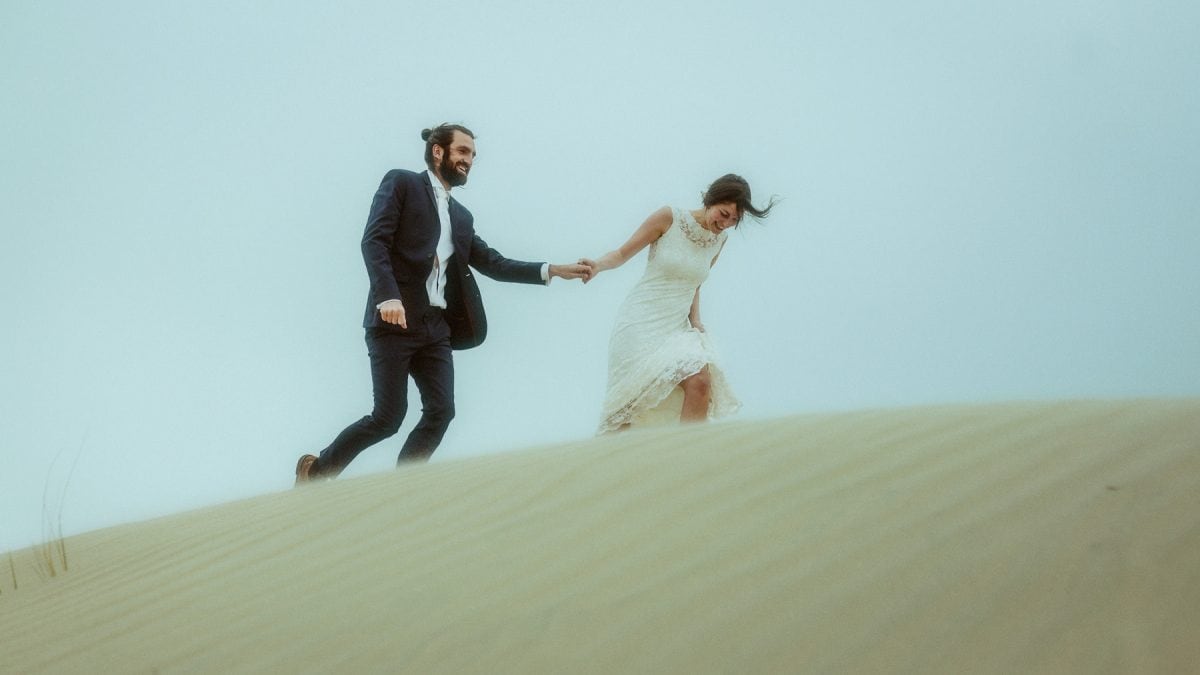 Couple in wedding attire running hand in hand over the windswept sand dunes of Rømø, Denmark—her lace dress flowing, his suit jacket catching the breeze. A candid moment of laughter, movement, and wild North Sea energy.