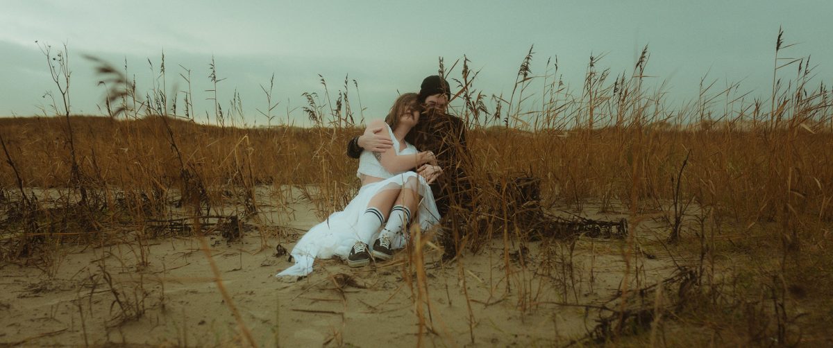 Newlyweds sitting close in the windswept dunes of Rømø, Denmark—her white dress gathered around her, his arms wrapped protectively, both lost in quiet laughter among golden sea grass under a moody sky. Raw, real, and wildly intimate.