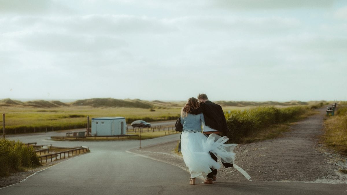 Ein Paar geht Arm in Arm eine leere Straße entlang, nahe St. Peter-Ording. Das Kleid und der Schleier der Braut wehen im Küstenwind, umgeben von Dünen und salziger Luft – ein stiller, intimer Moment voller Freiheit und Verbundenheit.