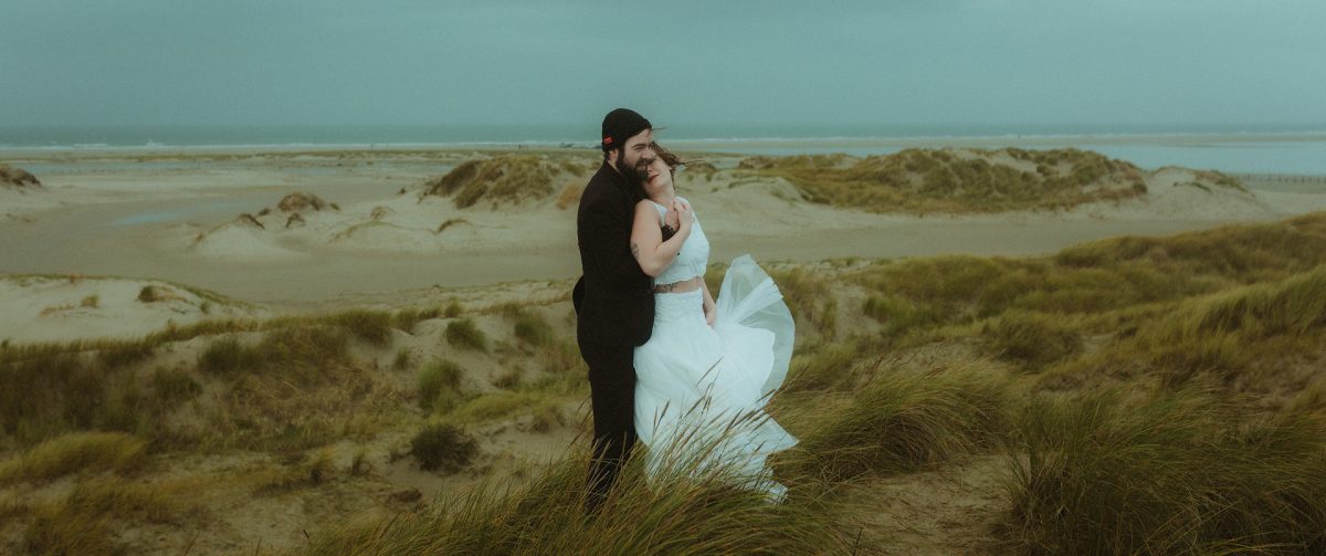 Elopement on Rømø: A couple embraces on a windswept dune overlooking the wild North Sea. Her dress dances in the stormy breeze—intimate, real, and full of raw North Sea vibes.. Perfect setting for an elopement at the North Sea.