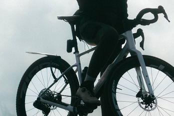 Cyclist in full black kit standing over a white Orbea Terra with Zipp wheels against a pale overcast sky, 2026