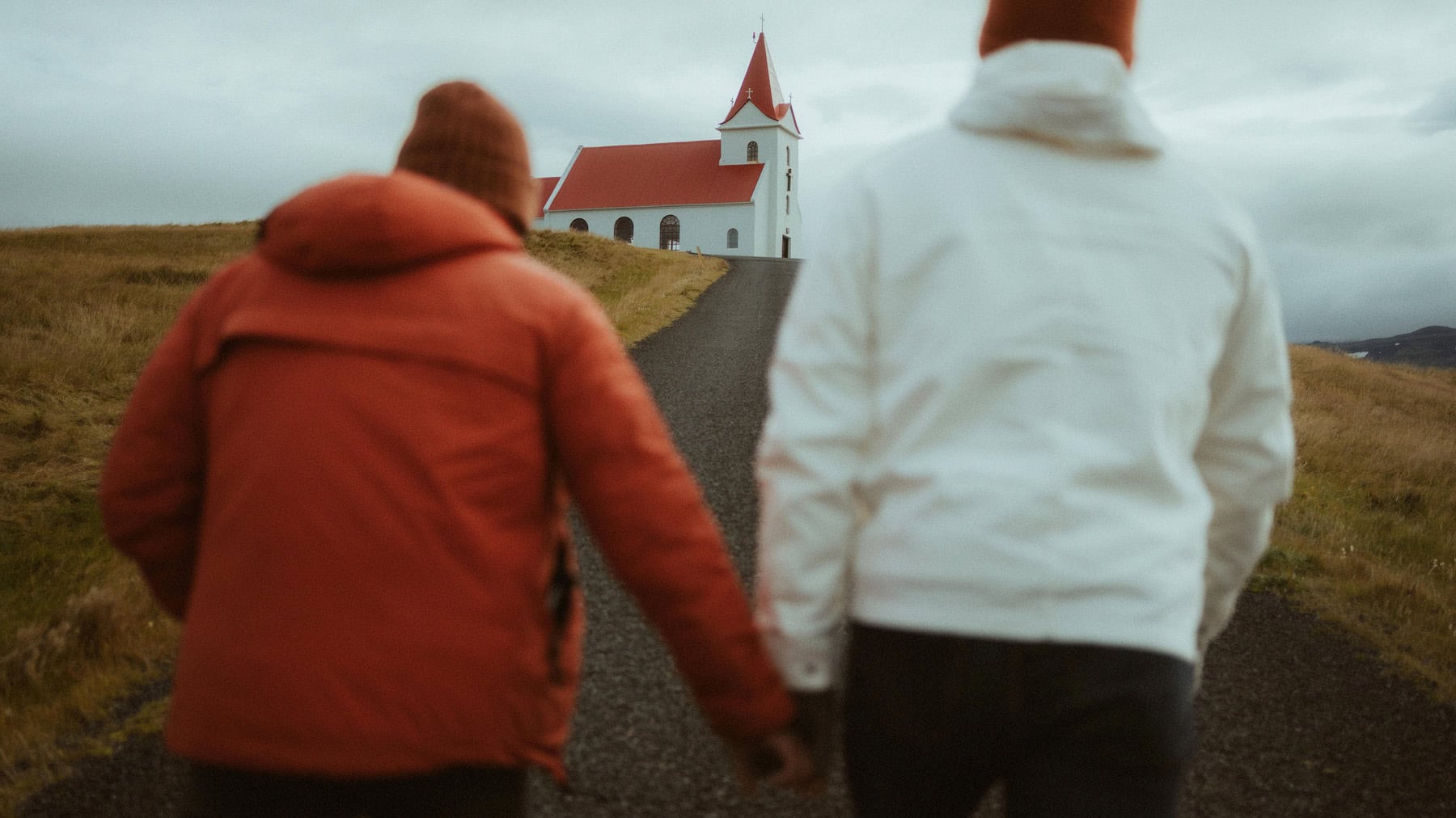 A couple walking up a gravel path toward a charming church with a red roof and white walls, perfectly capturing the intimate essence of an Iceland elopement, all set on a picturesque grassy hill.