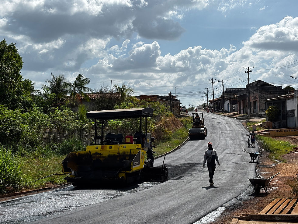 Obra de pavimentação em rua rural, com rolo compressor, operários e nuvens carregadas; placa ROUPA BOA E BARATA.