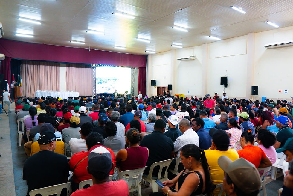 Auditório lotado com público sentado diante de palco e tela de projeção, em ambiente iluminado e silencioso.