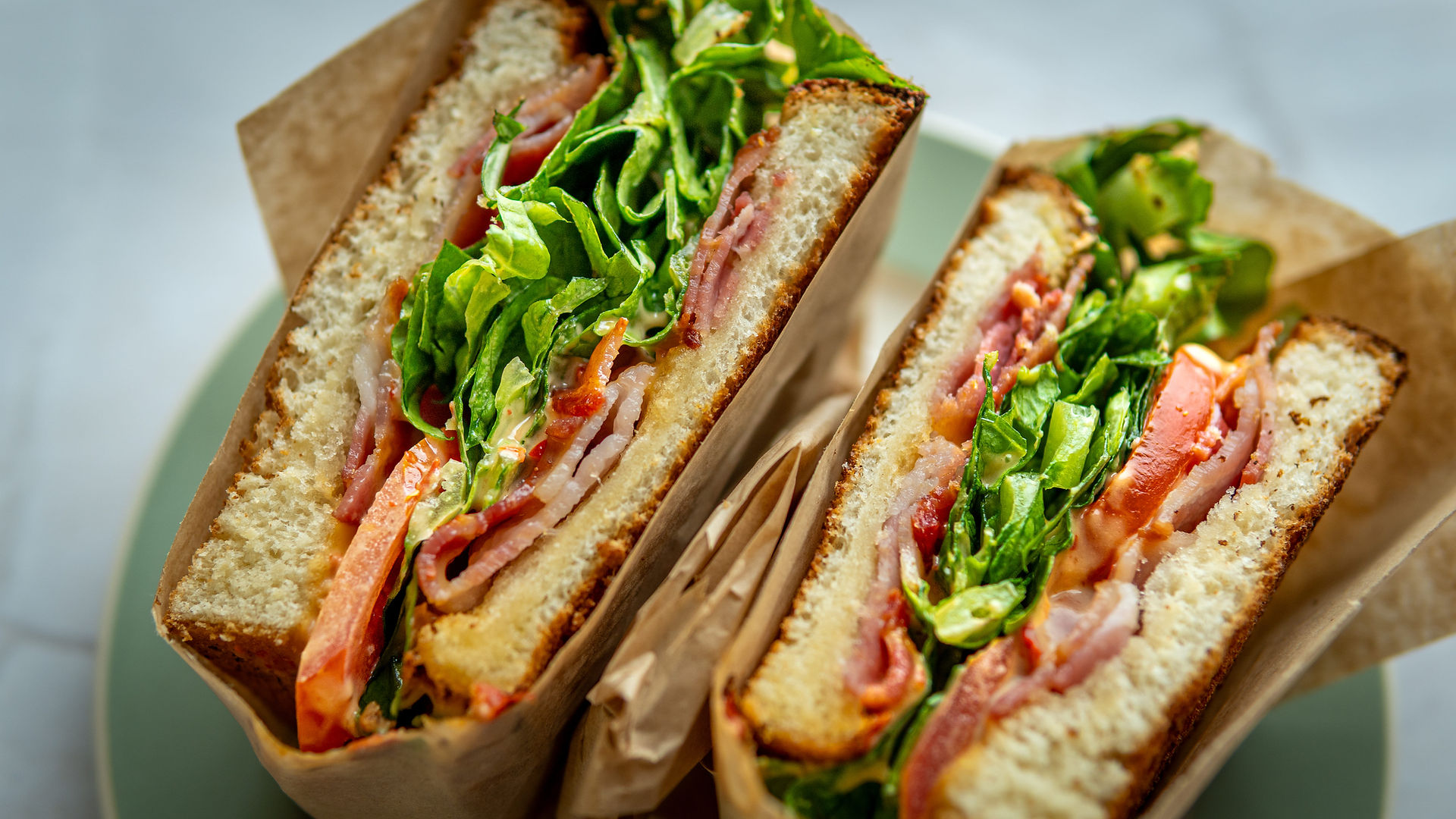 Een close-up van een versbereide BLT sandwich met knapperige bacon, verse sla en rijpe tomaten tussen twee dikke sneetjes goudbruin geroosterd brood. De sandwich is verpakt in bruin papier en ligt op een zachtgroen bord, wat de frisheid van de ingrediënten benadrukt.