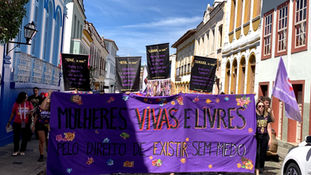 Pessoas marcham em rua de cidade histórica com cartazes com nomes das vítimas e uma faixa grande escrito: Mulheres Vivas e Livres. Pelo direito de Existir sem medo.