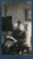 A black and white photo of a young man, sitting, legs crossed, on a high-back armchair. He stares directly into the camera, holding an opened book. Light streams in from the right of the frame and two framed pictures hang next to, and behind him. He is dressed in a suit and his face is expressionless.
