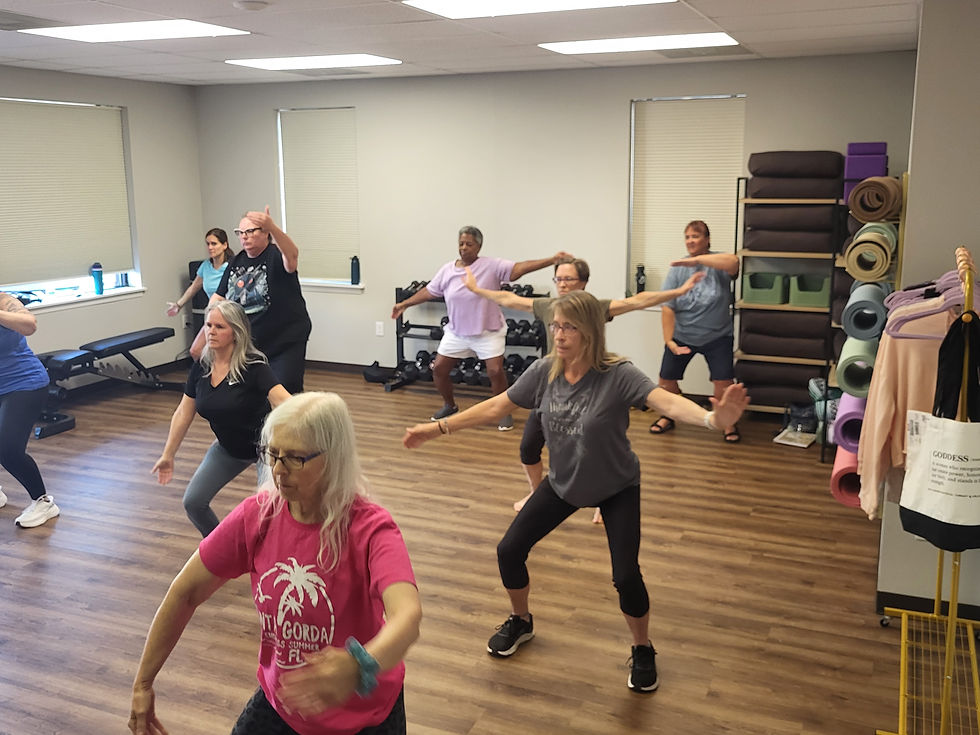 Adults in a group exercise class performing synchronized movements on a wood floor during Tai Chi Fustion class with Jane Hamann. Rolled yoga mats are stacked in the background.