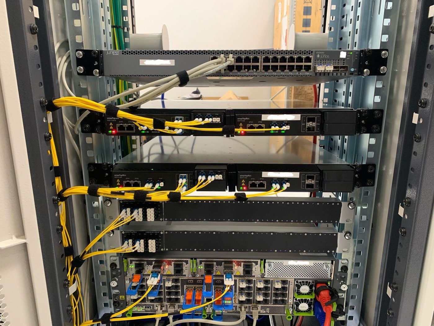Rack in Cape Town with a Juniper Ethernet switch, Smartoptics optical line systems and mux/demux, and a Juniper ACX7300 router with coherent DWDM plugs