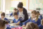 Young female teacher standing behind a male grade school student amidst a classroom of uniformed students working at their desks