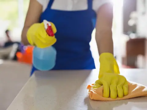Body shot of female wearing a blue apron, and yellow gloves while using cleaning products to disinfect a countertop