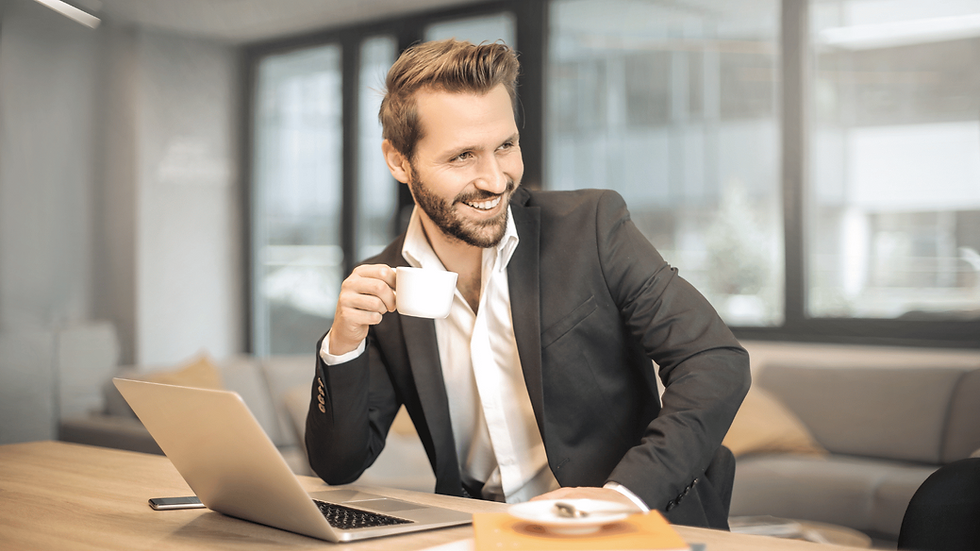 Man drinking an expresso and smiling at someone out of shot, while sitting at his desk, with laptop open.
