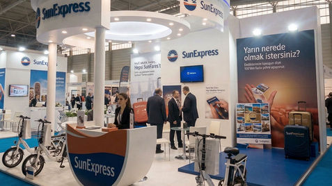 A modern stand designed by BayZ Design Architecture for the SunExpress brand at the international tourism fair, featuring curved lines in the shape of an aircraft fuselage, vibrant orange and blue corporate colors, and a digital check-in experience area.
