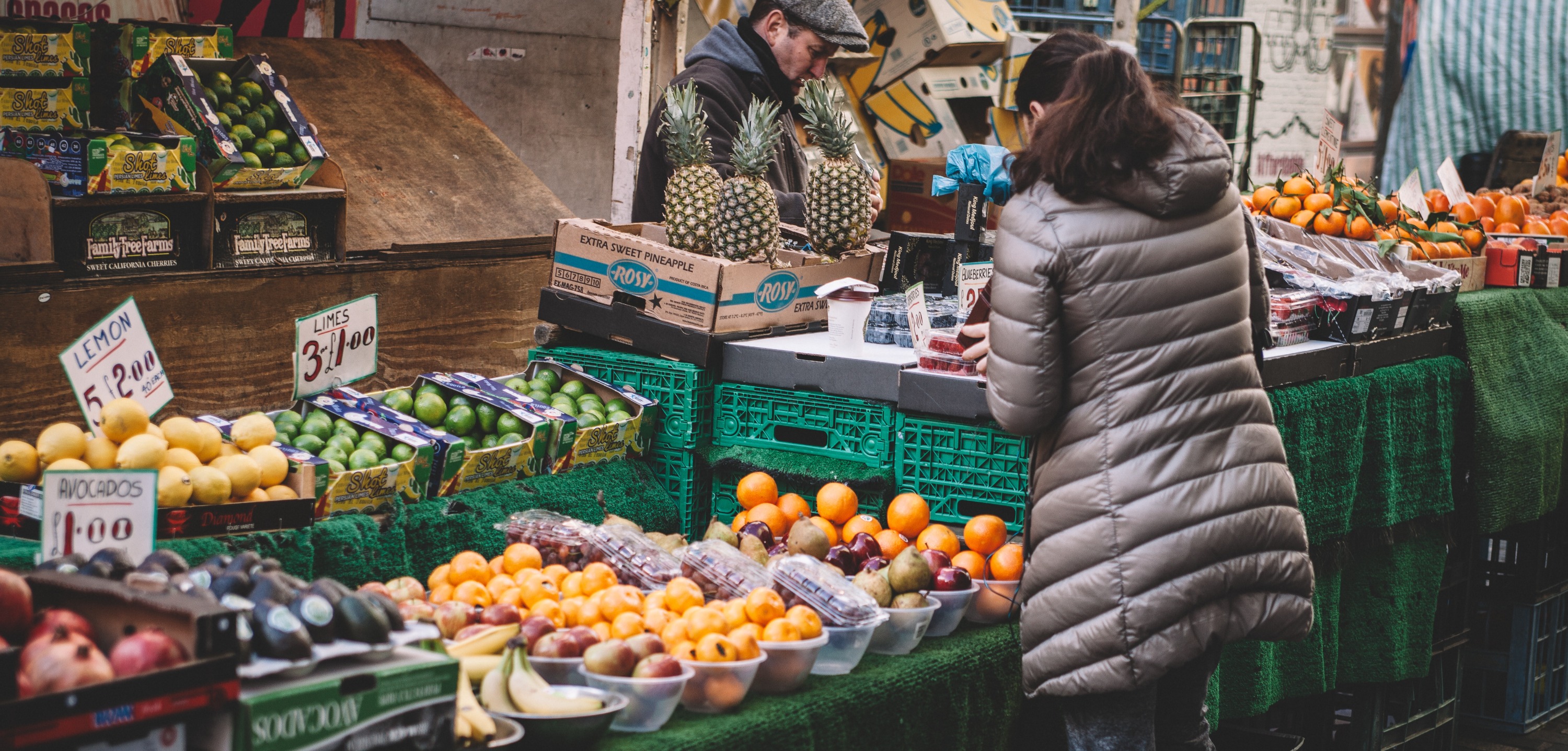 Puesto de frutas y verduras en un mercado al aire libre con clientes seleccionando productos, perfecto para quienes buscan darse de alta como autónoma en el comercio local.
