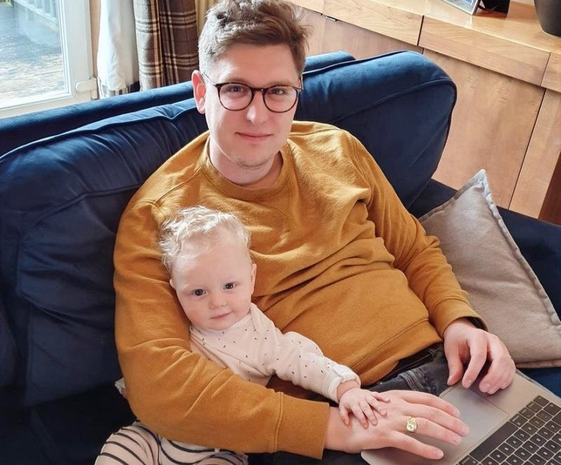 A man sits with his young son on a couch while working on his laptop