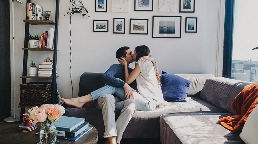 Facetime virtual photo of an anglo-american couple kissing. Woman is holding man's face while legs are intertwined with his as they are sitting on an L-shaped gray sofa in their home with light pouring in on their right through a large window and a wall full of framed photos as their background, a round desk with pink florals in the foreground and a bookcase on their left, taken by Carole Cohen Photography