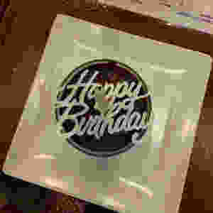 A white candle over a cursive "Happy Birthday" stencil over a chocolate mousse