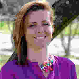 This is a headshot of Maura Horton. She is looking directly at the camera and smiling. She has light-brown hair to her shoulders and she is wearing a purple blouse and statement necklace that has many different colors including turquoise blue, green, orange, pink, and purple. She is in front of a tree.