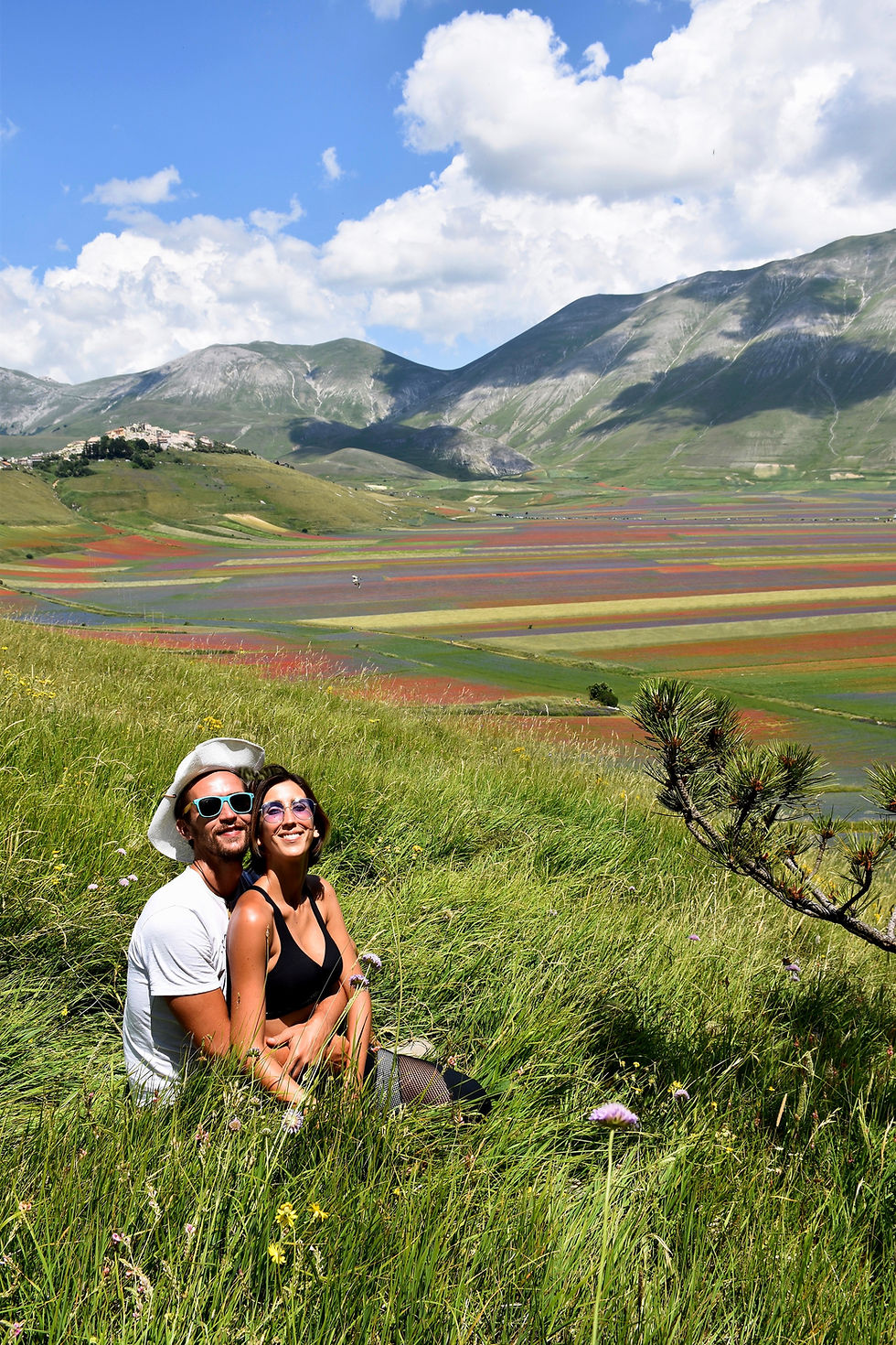 Eugenia e Nicola abbracciati, seduti sul prato verde, e alle nostre spalle la colorata fioritura di Pian Grande e Castelluccio di Norcia, Umbria.
