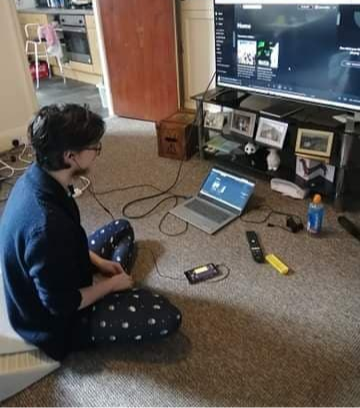 A picture of a man with dark hair wearing a navy blue cardigan and pyjama bottoms with black earphones plugged into a black smartphone. On the phone the app is visible. Paralell to the man, a grey laptop is open and displaying spotify with a green witch with black hat to signify the Wizard of Oz soundtrack. On the ground beside the man is a remote control and bottle of juice.
