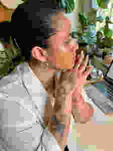 Author Karla Cornejo Villavicencio sitting at a desk with a computer, hands held together, the author stares into distance, surrounded by plants