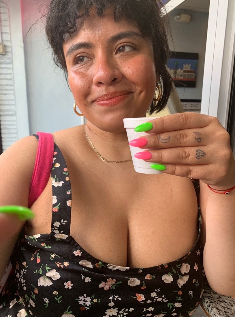 The author smiles while holding a small white cup with long pink and green nails