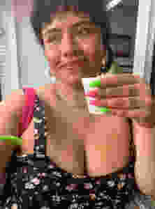 The author smiles while holding a small white cup with long pink and green nails