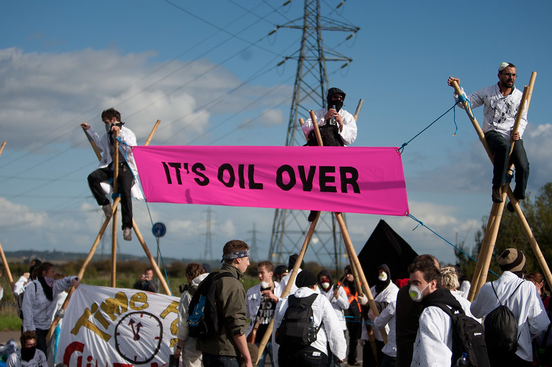 Three people sitting on tripods above a crowd holding a pink banner saying "IT'S OIL OVER"