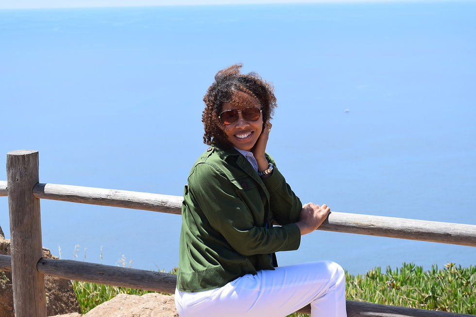 Lauren Francis-Sharma , the author, sits on an overlook of ocean, looking at camera and holding her hair back from the wind