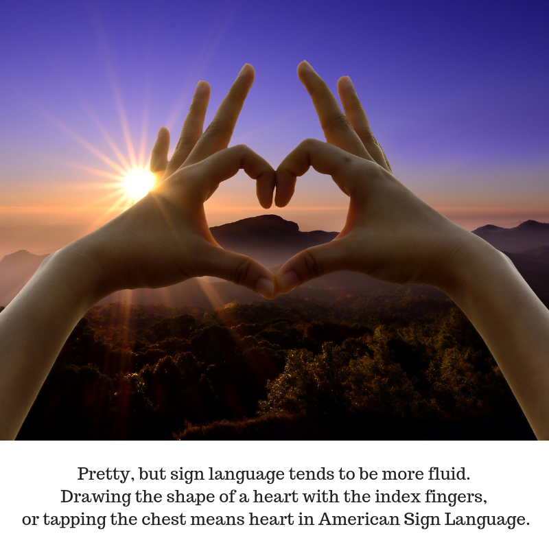 Close up of a pair of hands with the fingers in the shape of a heart against a vivid sunlit mountainous landscape.