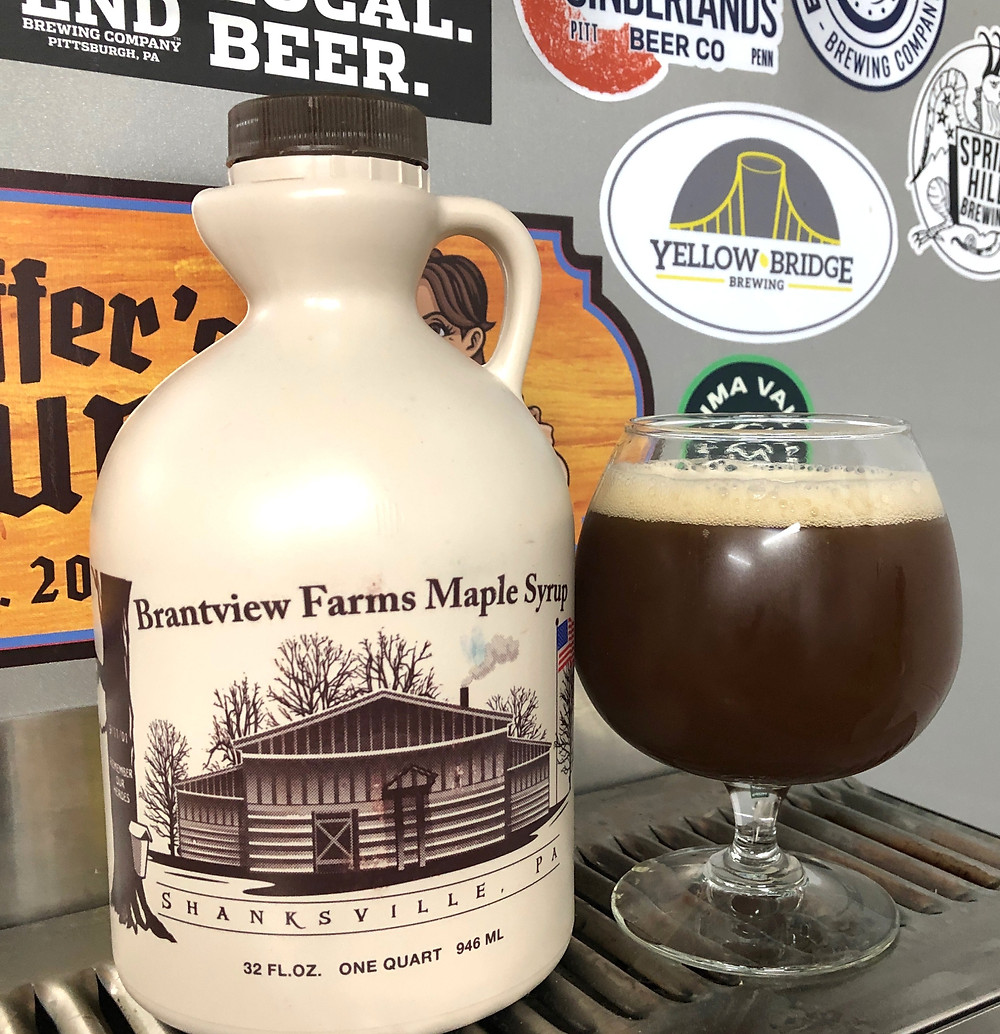 A jug of Brantview Farms Maple syrup sits next to a glass filled with a brown beverage that has a light tan foam on top. The jug and glass are resting atop a silver grate of a beer tap with various colorful stickers on wall behind them.