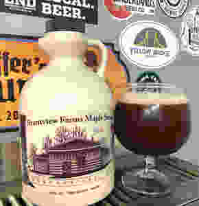 A jug of Brantview Farms Maple syrup sits next to a glass filled with a brown beverage that has a light tan foam on top. The jug and glass are resting atop a silver grate of a beer tap with various colorful stickers on wall behind them.