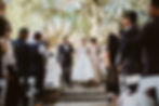 A bride walks down the aisle with the mother of the bride and the father of the bride by her side. They escort her to the groom waiting at the alter under an arch in an outdoor arena surrounded by large trees. The wedding planner fluffed her dress right before she walked onto the outdoor patio for the couple's wedding ceremony.