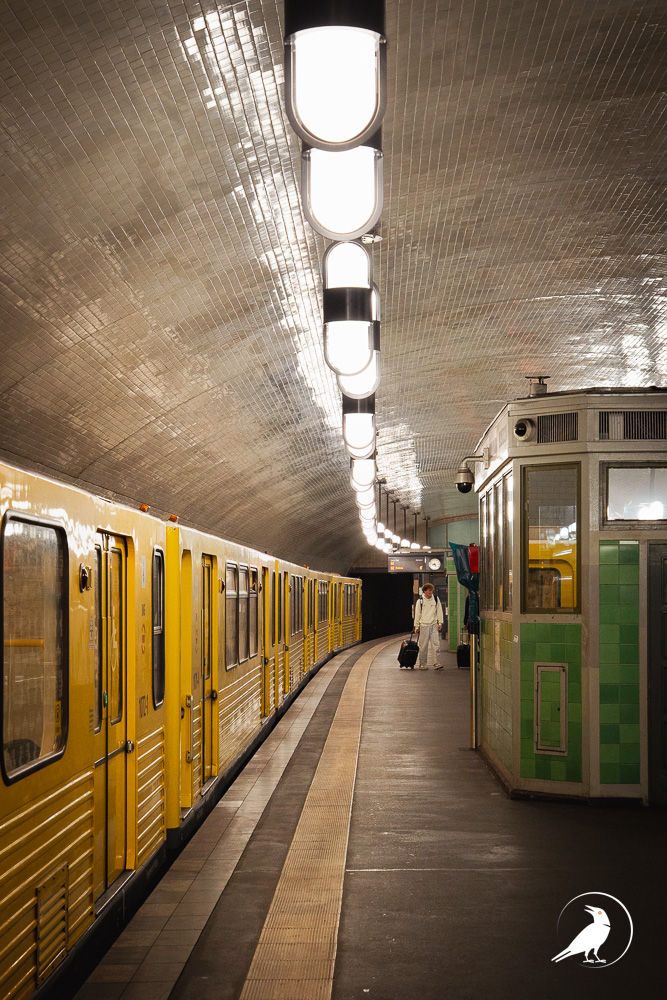 Yellow subway train in a tiled underground station; a lone traveler pulls a suitcase under bright ceiling lights.