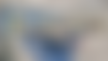 Modern school common area with collaborative seating, featuring light wood tables surrounded by blue plastic chairs on a multicolored floor. In the background are soft lounge seats, lockers along the wall, a kitchenette with a sink and microwave, and an open doorway leading to a restroom. The space is brightly lit with recessed ceiling lights and designed for flexible student use.