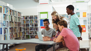 A group of students gathered around a laptop at a collaborative classroom table in a school library. Bookshelves line the background, and the image highlights teamwork, technology, and flexible learning spaces.