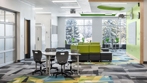 Modern collaborative classroom featuring flexible tables, rolling chairs, soft seating, and breakout spaces. Large windows provide natural light, while green accents, writable surfaces, and varied seating areas support group work, discussion, and student engagement.