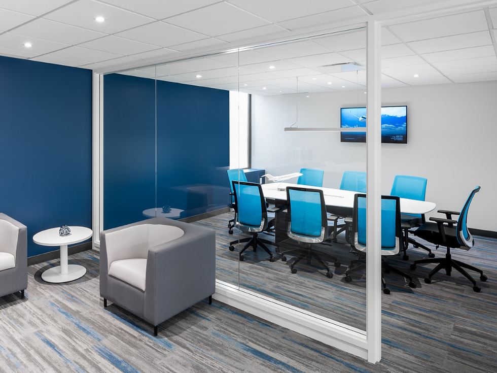 A modern office meeting space featuring a glass-enclosed conference room with a white meeting table surrounded by blue ergonomic office chairs. Outside the meeting room, two gray lounge chairs and a small round side table create a comfortable waiting area. Blue accent walls, patterned carpet, recessed lighting, and a wall-mounted display contribute to the clean, contemporary workspace design.