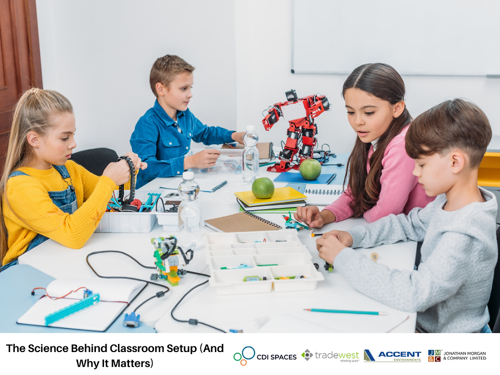 Four children sit around a classroom table building and programming small robots with electronic components and tools. A larger red robot model stands in the center of the table, while notebooks, water bottles, and green apples are scattered around.