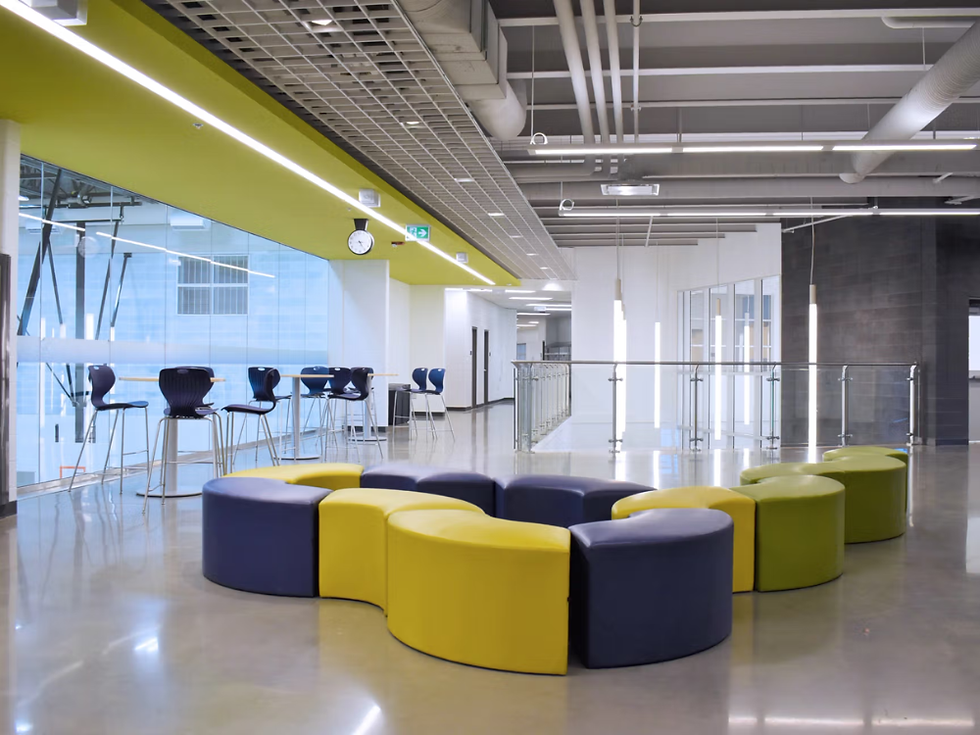 Open, modern school common area with polished floors, high exposed ceilings, and bright accent walls. Curved modular seating in blue, yellow, and green forms a circular arrangement in the center, while high-top tables and chairs line a glass wall, creating a collaborative and flexible gathering space.