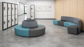 Modern school hallway featuring modular lounge seating in gray and teal, lockers, and wood-paneled walls. Circular and curved seating arrangements create collaborative student spaces in a bright, open learning environment.