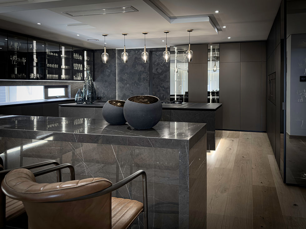 Modern kitchen with sleek gray marble countertops, pendant lights, and glassware cabinets. Brown leather chairs add warmth to the elegant setting.