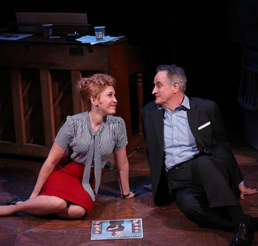 Performer in a gray blouse and red skirt seated on the floor beside a man in a dark suit, Smart Blonde