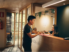 Reception staff member standing calmly behind a service counter in a health or community services setting, viewed from the side, representing occupational violence and aggression (OVA) and psychological safety in the workplace.