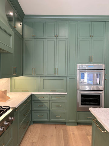 A corner of a kitchen is shown, featuring an abundance of sage green cabinetry, complemented by sleek gold hardware and a built-in stainless steel double oven.  White marble countertops provide a clean contrast to the cabinetry, creating a sophisticated and modern aesthetic.