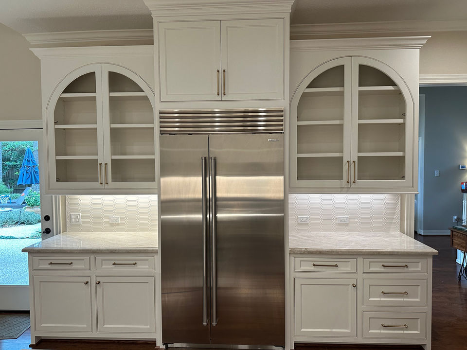 A stainless steel, built-in refrigerator is flanked by matching white cabinets, each with arched glass doors above and drawers below;  the counter-top is a light grey marble.