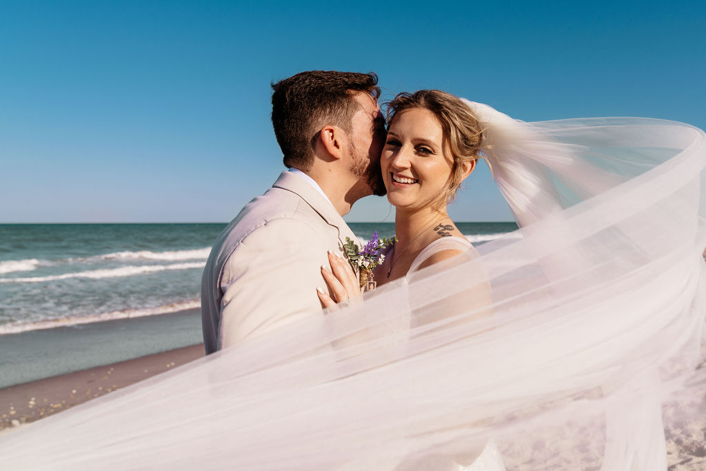 Bride smiling at the groom as her veil blows across their beach portrait