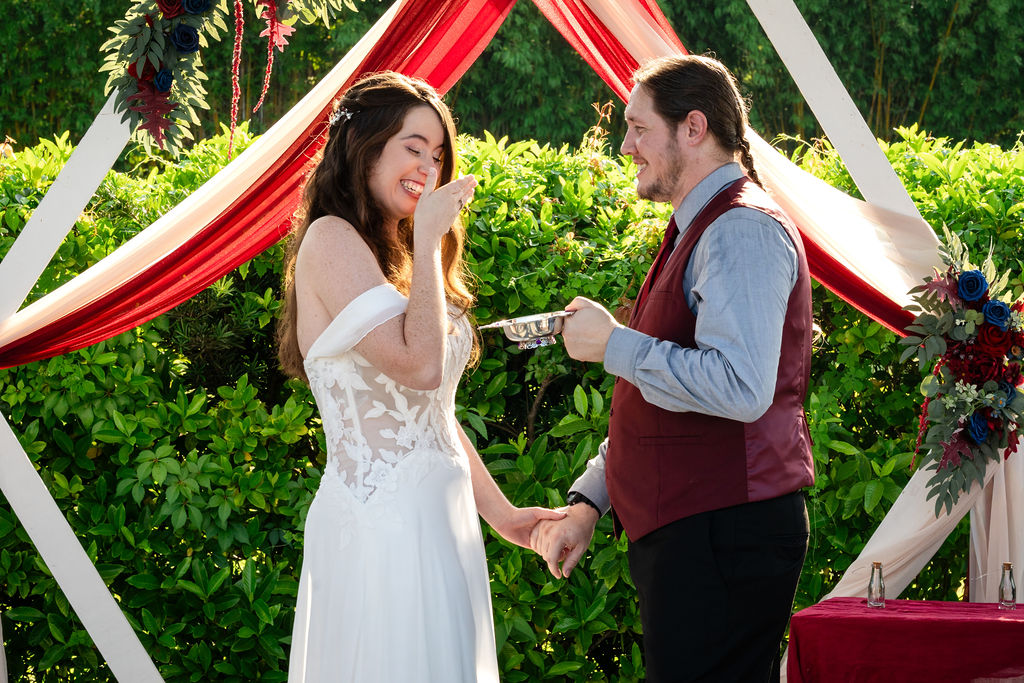 Bride and groom exchanging rings beneath a red-draped wedding arch